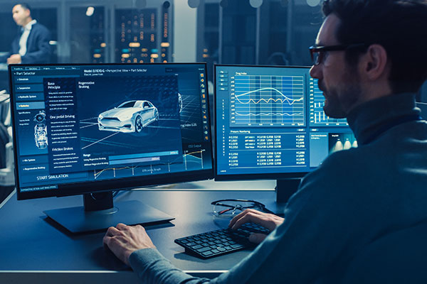 Professional Engineer Works on a Computer with a 3D CAD Software and Tests the Electric Car Chassis Prototype with Wheels, Batteries and Engine Standing in a High Tech Development Laboratory