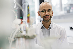 A male scientist is portrayed inside a laboratory. He's wearing glasses and looking friendly into the camera.